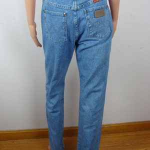 May include: Light blue denim jeans with a classic five-pocket design. Features copper rivets, a brown leather patch with the brand name, and a red logo tag. The jeans have a straight leg cut.