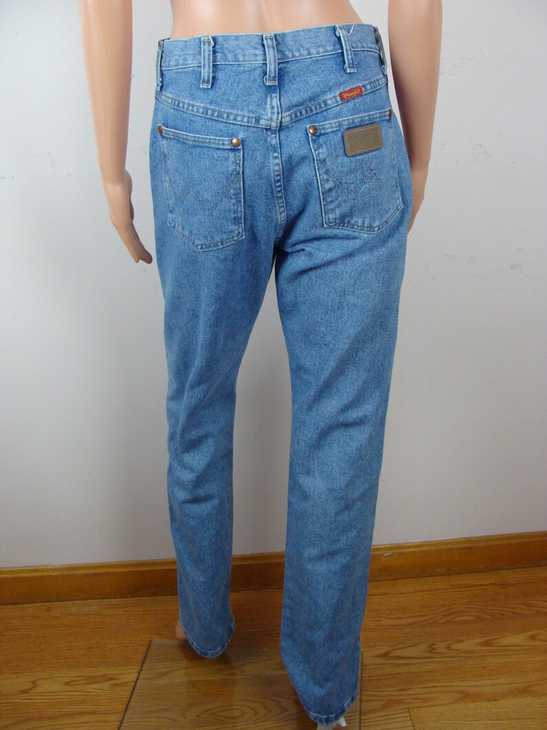 May include: Light blue denim jeans with a classic five-pocket design. Features copper rivets, a brown leather patch with the brand name, and a red logo tag. The jeans have a straight leg cut.