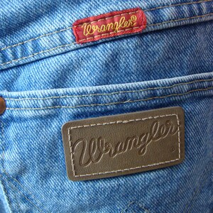May include: Close-up of light blue denim jeans with two "Wrangler" brand labels. One label is red with gold lettering, and the other is brown with embossed lettering. The jeans have visible stitching and a classic pocket design.