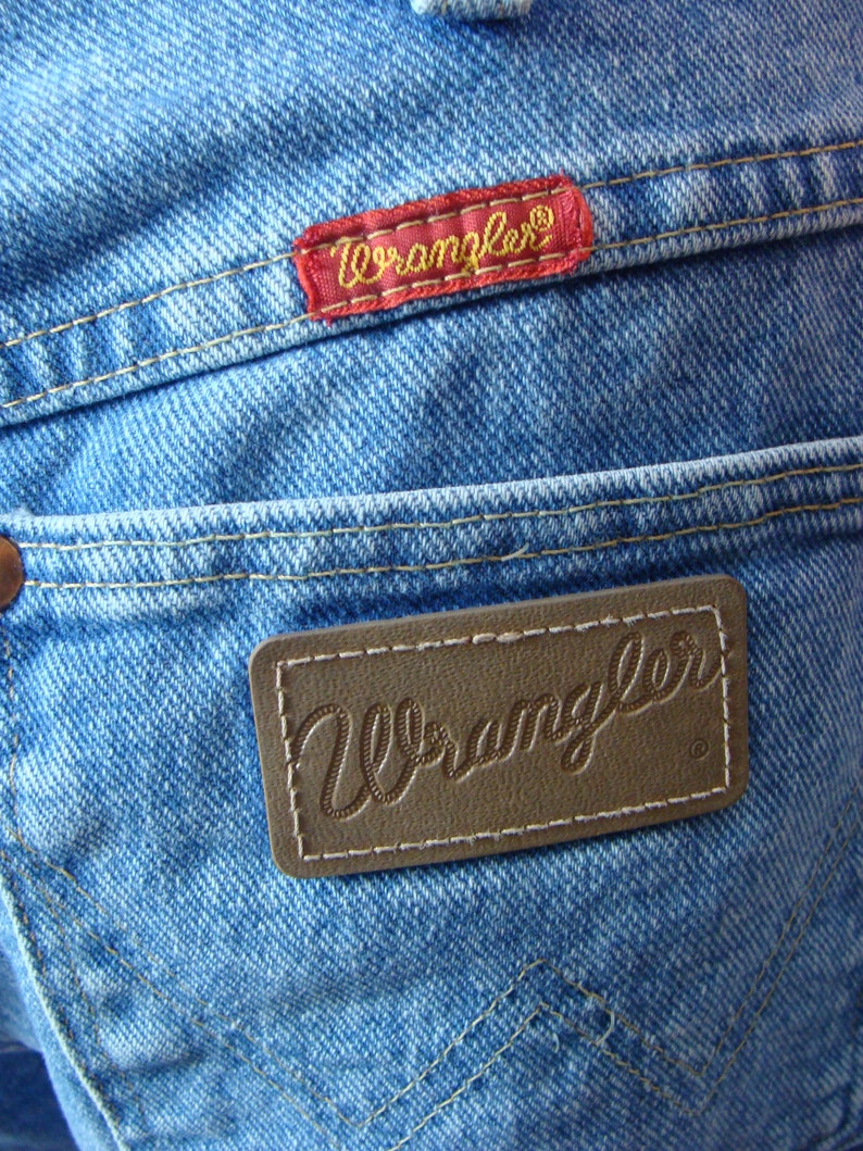 May include: Close-up of light blue denim jeans with two "Wrangler" brand labels. One label is red with gold lettering, and the other is brown with embossed lettering. The jeans have visible stitching and a classic pocket design.