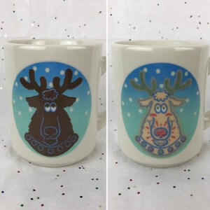May include: Two white ceramic mugs with a blue and green background. Each mug features a cartoon reindeer with a red nose and brown antlers. The reindeer on the left mug is brown and the reindeer on the right mug is beige.