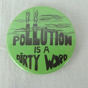 May include: Green button with black text that reads "Pollution is a dirty word". The button features a cartoon image of two smokestacks with smoke coming out of them.