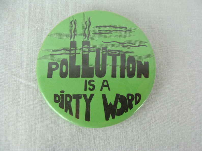 May include: Green button with black text that reads "Pollution is a dirty word". The button features a cartoon image of two smokestacks with smoke coming out of them.