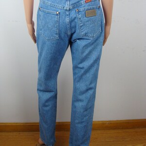 May include: Light blue denim jeans with a brown leather belt. The jeans have two back pockets and a Wrangler brand patch. The belt has a decorative cut-out design. The jeans are displayed on a mannequin.