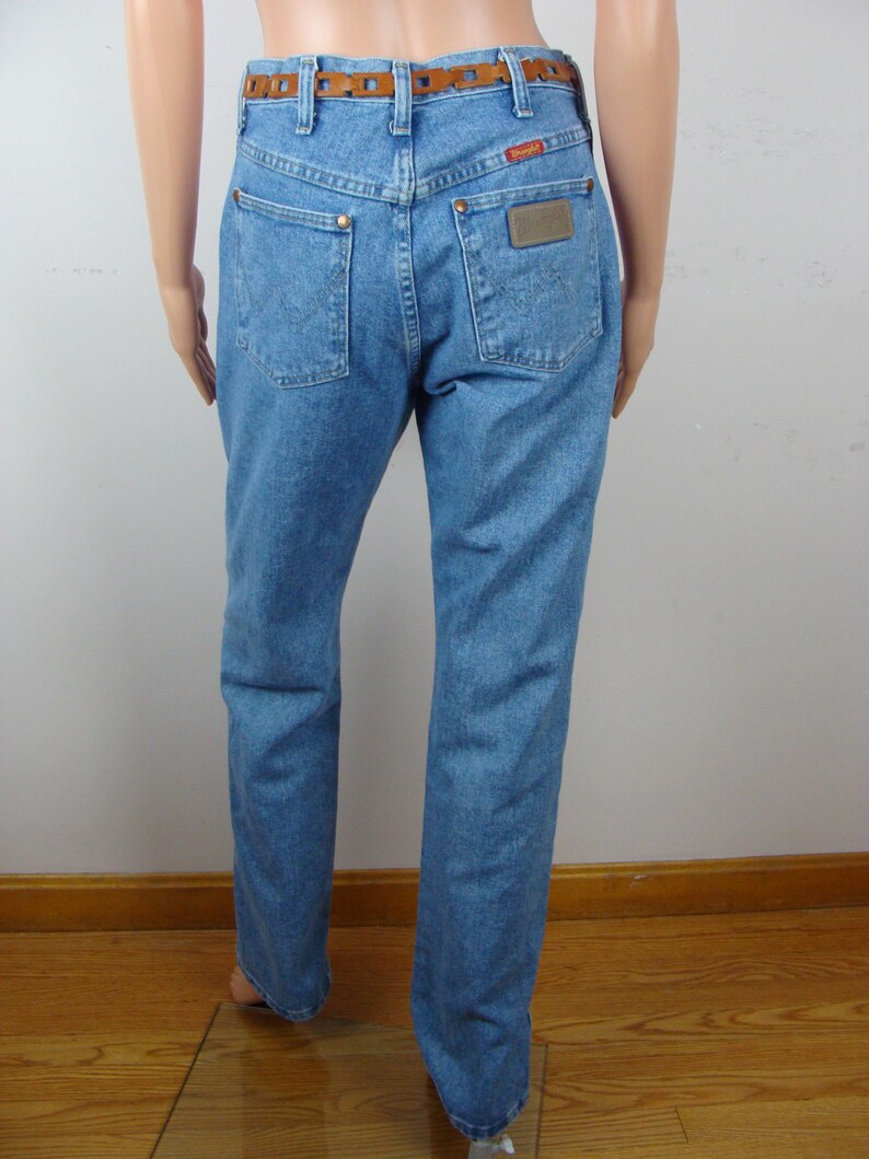 May include: Light blue denim jeans with a brown leather belt. The jeans have two back pockets and a Wrangler brand patch. The belt has a decorative cut-out design. The jeans are displayed on a mannequin.