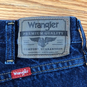 Op de afbeelding: Een blauwe denim jeans met een bruin leren patch waarop "Wrangler Premium Quality" staat met een adelaarlogo en de tekst "EST. 1947 COMFORT. GUARANTEED. GREENSBORO, NORTH CAROLINA". Onder de bruine patch zit een rode leren patch met de tekst "Wrangler".