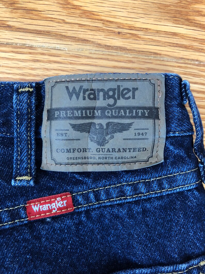 Op de afbeelding: Een blauwe denim jeans met een bruin leren patch waarop "Wrangler Premium Quality" staat met een adelaarlogo en de tekst "EST. 1947 COMFORT. GUARANTEED. GREENSBORO, NORTH CAROLINA". Onder de bruine patch zit een rode leren patch met de tekst "Wrangler".