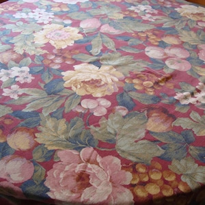May include: Round tablecloth with a floral pattern. The fabric features large pink, yellow, and white flowers, green leaves, and clusters of grapes against a burgundy background. The tablecloth is on a wooden table.
