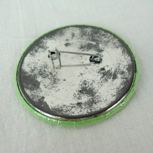 May include: The back of a green button with a metal pin. The pin is attached to the button with a metal loop.