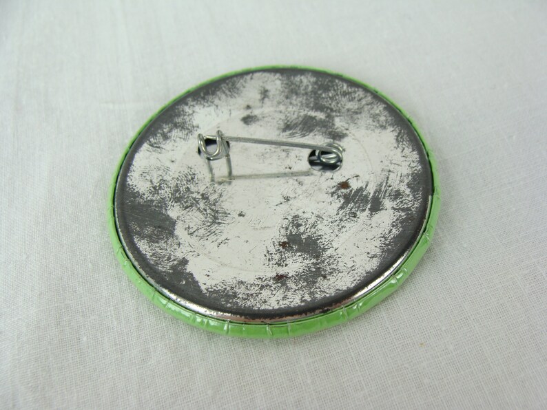 May include: The back of a green button with a metal pin. The pin is attached to the button with a metal loop.