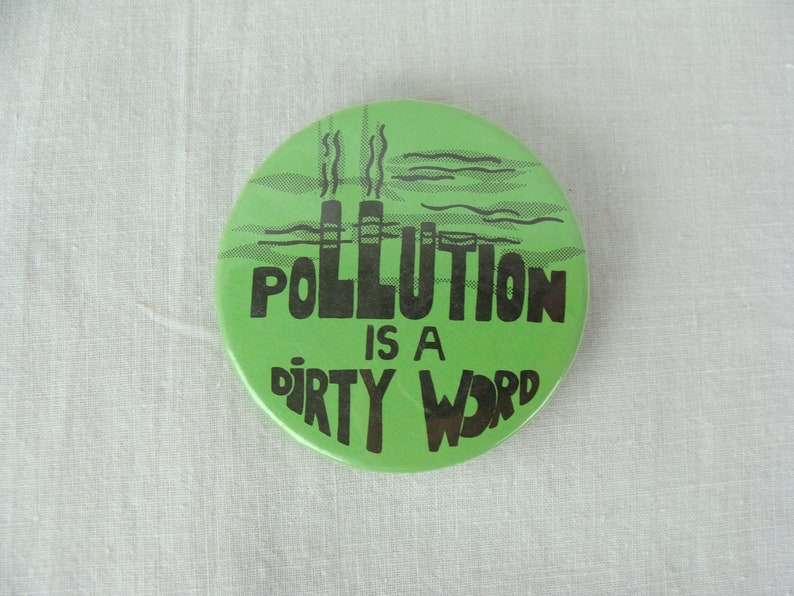 May include: A green button with black text that reads "Pollution is a dirty word". The button features a black silhouette of factory smokestacks and smoke.