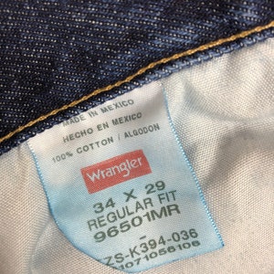 Op de afbeelding: Een wit label met zwarte tekst die "MADE IN MEXICO", "HECHO EN MEXICO", "100% COTTON / ALGODON", "Wrangler", "34 X 29", "REGULAR FIT", "96501MR", "ZS-K394-036", "1071056106" luidt.