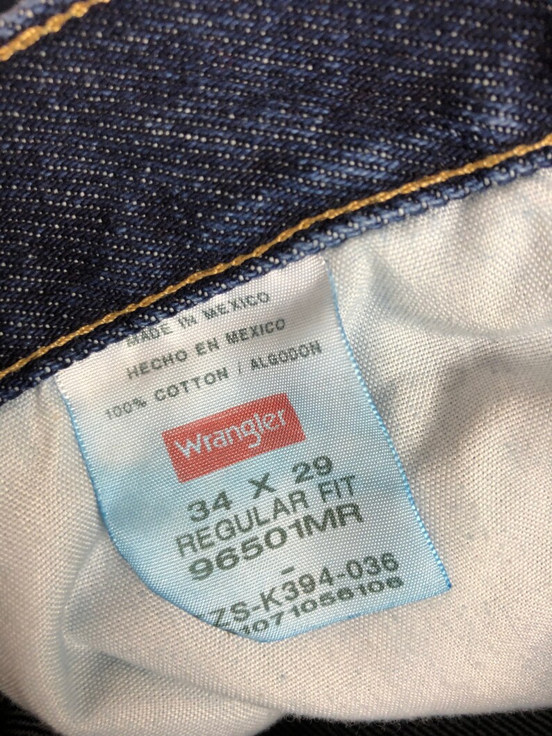 Op de afbeelding: Een wit label met zwarte tekst die "MADE IN MEXICO", "HECHO EN MEXICO", "100% COTTON / ALGODON", "Wrangler", "34 X 29", "REGULAR FIT", "96501MR", "ZS-K394-036", "1071056106" luidt.