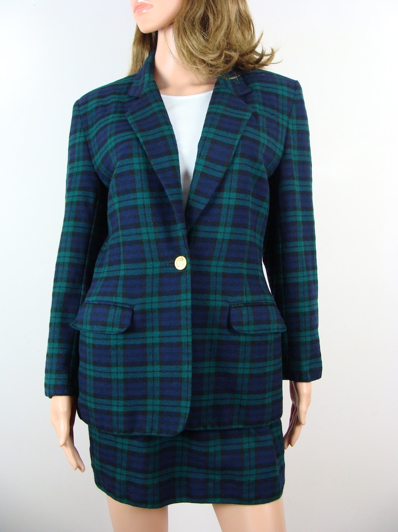 Op de afbeelding: Een groen en blauw geruit blazer- en rokpak. De blazer heeft een enkele knoopsluiting en een zak aan de linkerkant. De rok is knielengte.