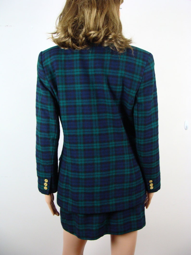 Op de afbeelding: Een blauwe en groene geruite blazer en rok set. De blazer heeft gouden knopen aan de manchetten. De rok is een minirok.