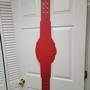 May include: A red, decorative belt-shaped item with silver rivets, hanging on a white door. The item has a central, rounded shape and a long, rectangular extension on both ends, each with multiple rivets.