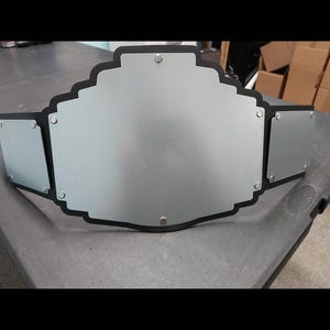 May include: A silver-colored championship belt with a black border. The belt has a large central plate and two smaller side plates, all with rounded edges. The plates are attached to a black strap.