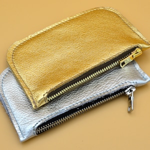 Puede incluir: Dos estuches con cremallera, uno dorado y otro plateado, están apilados uno encima del otro. El estuche dorado está en la parte superior y el estuche plateado está en la parte inferior.