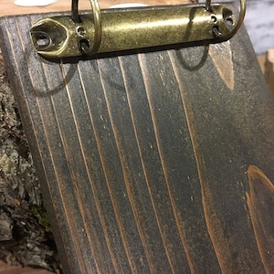 May include: A dark stained wooden clipboard with a brass-coloured binder clip. The wood grain is visible, and the clipboard is angled, showing the texture and colour variations. The binder clip has two rings.