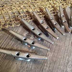 May include: A set of ten silver metal boat cleats, arranged in a semi-circle on a weathered wooden surface. The cleats are a nautical decor item.