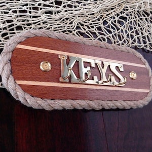 May include: Wooden key holder with a rope border and brass hooks. The holder has a light wood background with dark wood stripes and the word "KEYS" in brass letters.