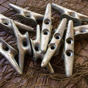 May include: A close-up of a group of silver metal cleat hooks, also known as boat cleats, arranged on a brown and tan net background. The cleats are in various positions and angles, showing their unique shape and design.