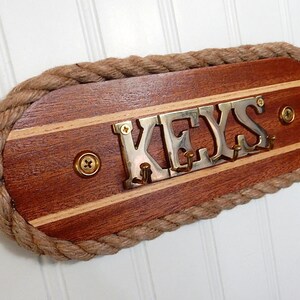 May include: A wooden key holder with a rope border. The holder has a natural wood finish with a light wood inlay. The word "KEYS" is spelled out in brass letters with hooks for hanging keys.