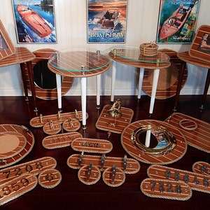May include: A collection of nautical themed home decor items, including tables, trays, and wall decor. The items are made of wood and have a rope trim. The tables have glass tops and are decorated with nautical symbols. The trays are round and have a cork centre. The wall decor includes a porthole mirror and a set of house numbers.