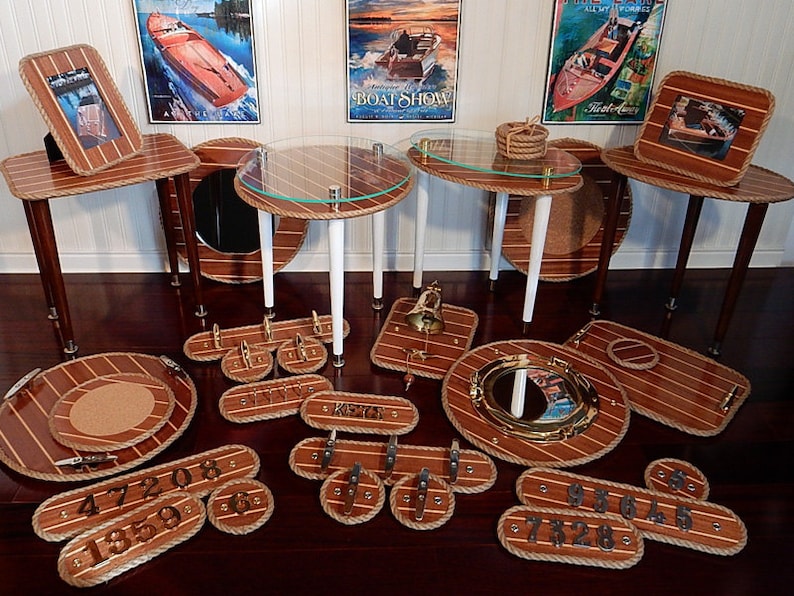 May include: A collection of nautical themed home decor items, including tables, trays, and wall decor. The items are made of wood and have a rope trim. The tables have glass tops and are decorated with nautical symbols. The trays are round and have a cork centre. The wall decor includes a porthole mirror and a set of house numbers.