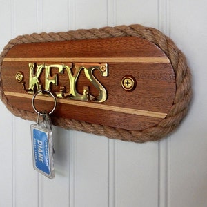 May include: A wooden key holder with a rope border. The holder has a brass plaque that says "KEYS" and a key ring with a key attached.