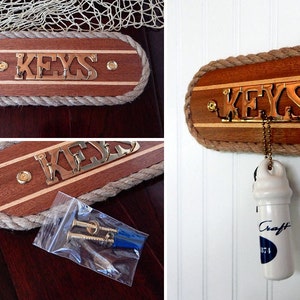 May include: A wooden key holder with a rope border and the word "KEYS" in brass letters. The holder is mounted on a white wall with a white ceramic bottle stopper hanging from it.