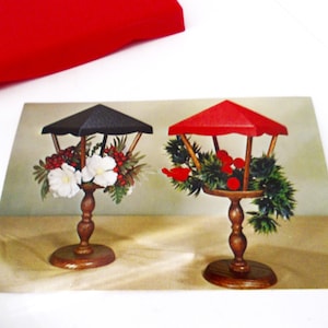 May include: Two vintage Christmas centerpieces with wooden bases and red and black fabric canopies. The centerpiece on the left has white flowers and red berries. The centerpiece on the right has green pine branches and red berries.