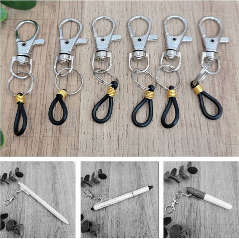 May include: Six silver keychains with black and gold accents. Each keychain has a silver ring and a black loop. The keychains are arranged in a row on a wooden surface.