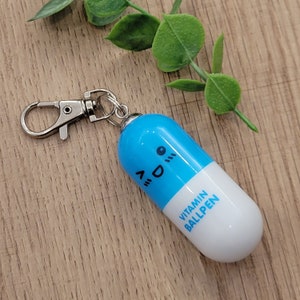 Blue mini pen shaped like a pill with emoji face attached to a silver clip to attach to badge reels, backpacks etc