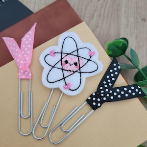 May include: Three decorative paper clips with ribbon accents. One clip has a pink polka dot ribbon, another features a white and black atom design, and the third has a black polka dot ribbon. The clips are made of silver metal.