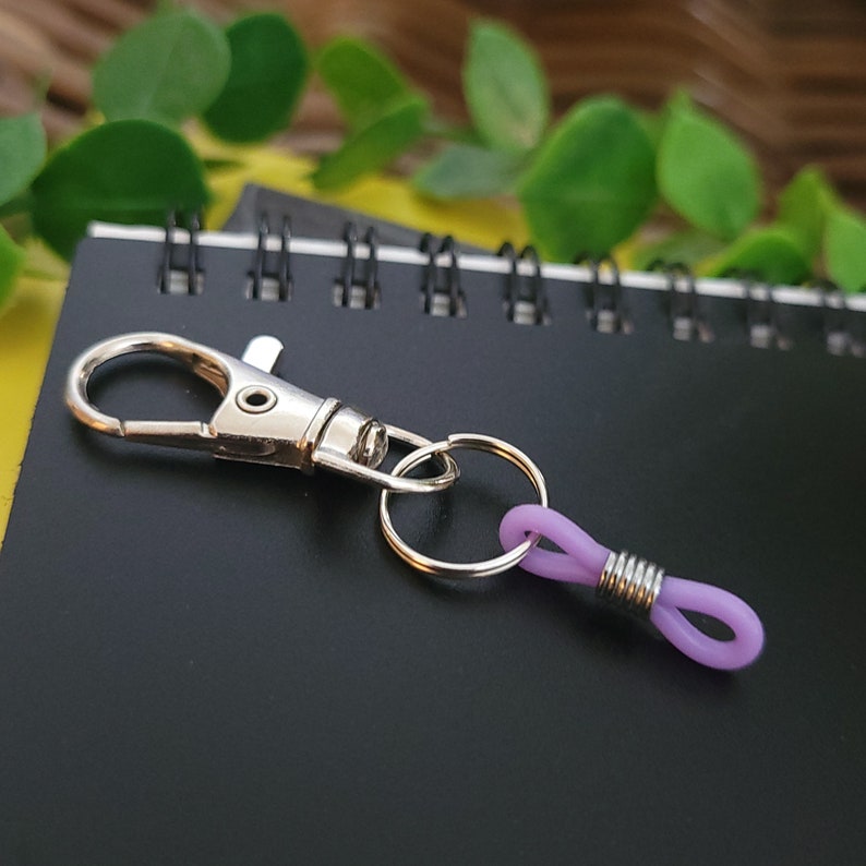 May include: A silver key chain with a purple rubber loop. The key chain has a silver ring and a silver hook.