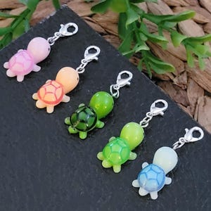 May include: Five colorful turtle charms with silver-tone lobster clasps. The turtles are in shades of pink, orange, green, and blue. Each charm has a detailed shell design and a glossy finish, perfect for jewelry making or crafting.
