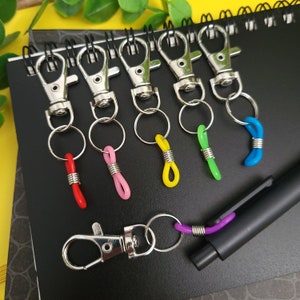 May include: Seven silver keychains with different colored plastic loops. The loops are red, pink, yellow, green, blue, and purple. The keychains are on a black surface.
