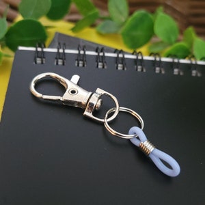 May include: A silver key chain with a silver loop and a light purple rubber loop. The key chain is lying on a black notebook with silver rings.
