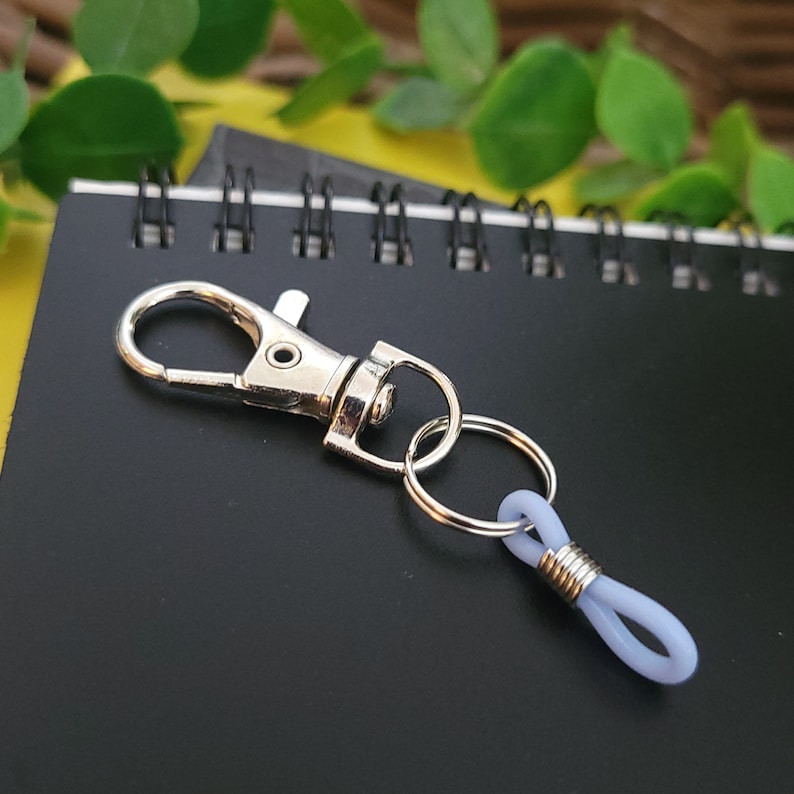 May include: A silver key chain with a silver loop and a light purple rubber loop. The key chain is lying on a black notebook with silver rings.