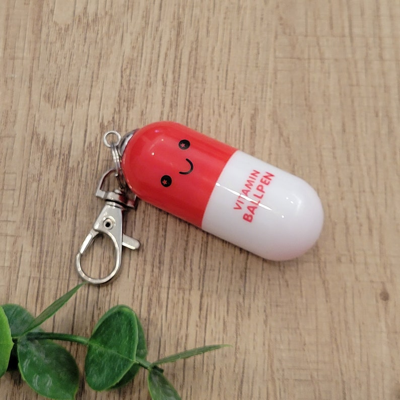 Red mini pen shaped like a pill with emoji face attached to a silver clip to attach to badge reels, backpacks etc