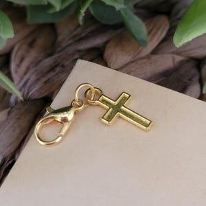 May include: A gold-colored charm featuring a cross pendant and a lobster clasp. The cross is a simple, classic design. The charm is likely made of metal and is shown against a neutral background.