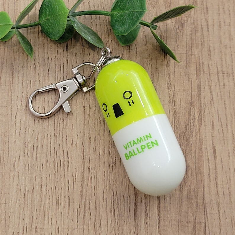 Lime Green mini pen shaped like a pill with emoji face attached to a silver clip to attach to badge reels, backpacks etc