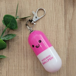 Hot Pink mini pen shaped like a pill with emoji face attached to a silver clip to attach to badge reels, backpacks etc