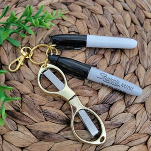 May include: A gold-colored keychain with a pair of small scissors and two Sharpie Mini markers. One marker is black, and the other is gray with the text "Sharpie MINI". The items are arranged on a woven, brown surface.
