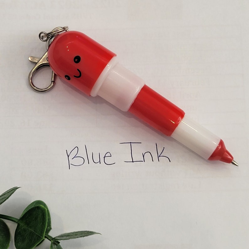 Red mini pen shaped like a pill with emoji face attached to a silver clip to attach to badge reels, backpacks etc