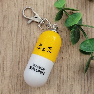 Yellow mini pen shaped like a pill with emoji face attached to a silver clip to attach to badge reels, backpacks etc