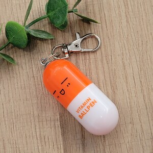 Orange mini pen shaped like a pill with emoji face attached to a silver clip to attach to badge reels, backpacks etc