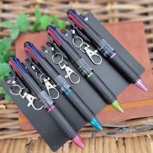 May include: Five multi-coloured retractable ballpoint pens with keychains attached. The pens are black, blue, red, green, and purple. The pens are on a brown leather surface.