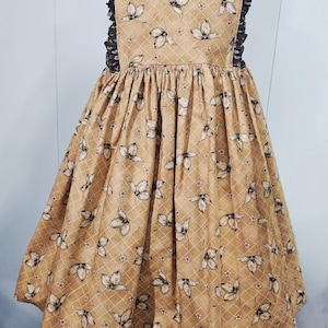 May include: A tan dress with a butterfly and floral pattern. The dress has a square neckline, ruffled black trim, and a gathered skirt. The fabric has a grid pattern with butterflies and small flowers. The dress is displayed on a wooden stand.